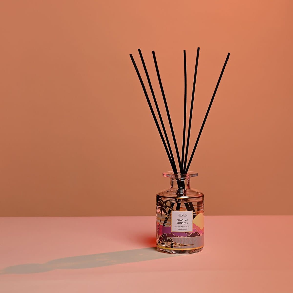 Glass reed diffuser bottle with wooden reeds beside a coastal sunset box