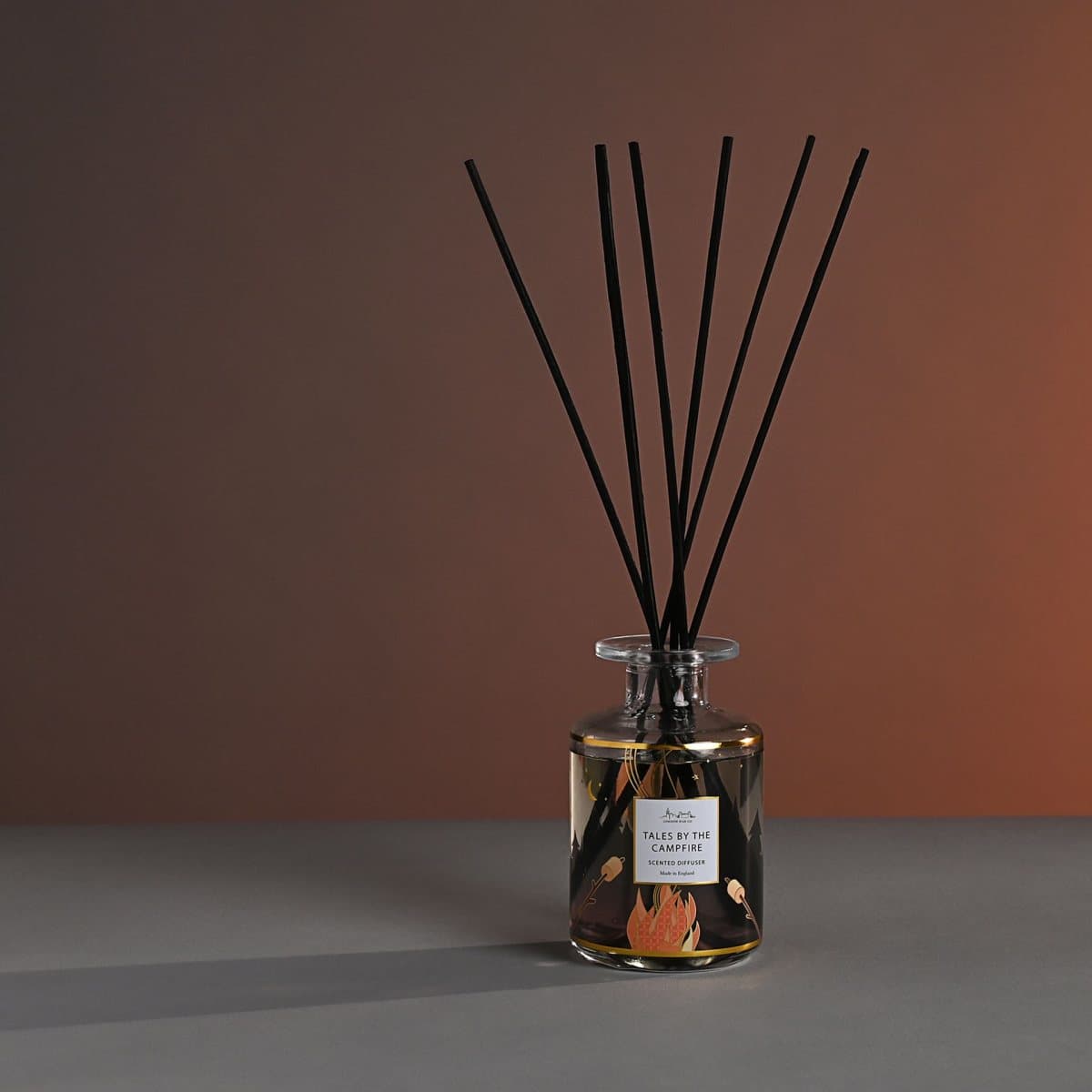 Glass reed diffuser bottle with wooden reeds beside a campfire design box